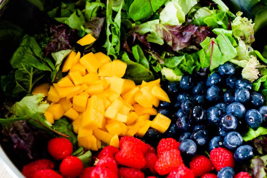 Bright, vibrant mangoes, raspberries and blueberries sit on top of a bowl full of dark mixed greens.