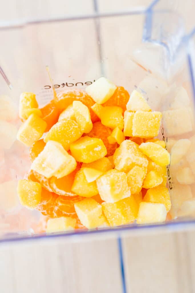 Looking down into a blender we see orange wedges and frozen mango chunks.