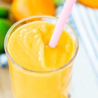 A tall glass of Orange Mango smoothie sits in front of oranges and limes. A pink straw sits in the glass.