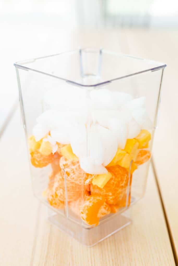 Blender sits on countertop filled with orange wedges, frozen mango chunks, lime juice, honey and ice cubes.