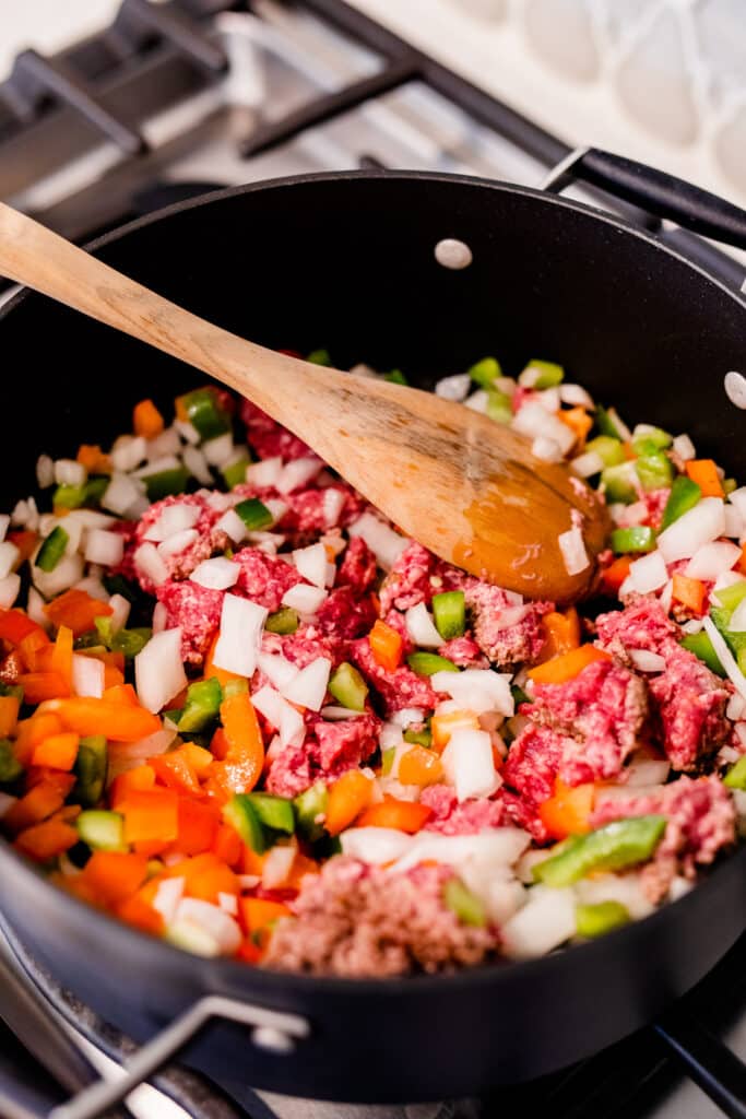 Ground beef, chopped onions, peppers and beans are stirred in a large black pot as they simmer and cook.