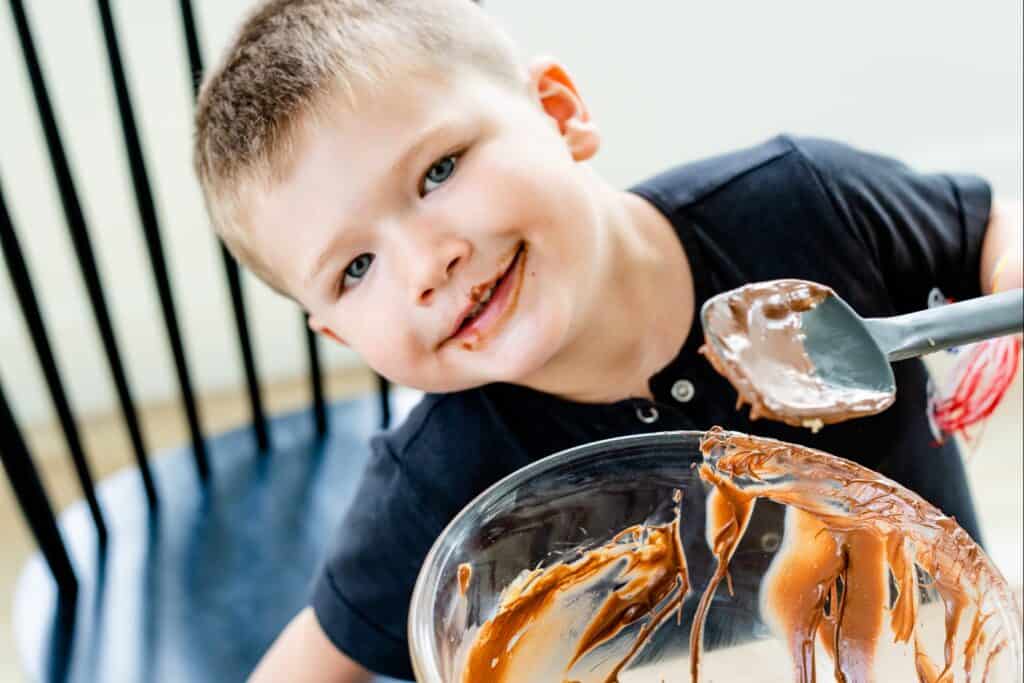 George sits at kitchen table with empty glass bowl with spatula in hand ready to clean off leftover chocolate topping.