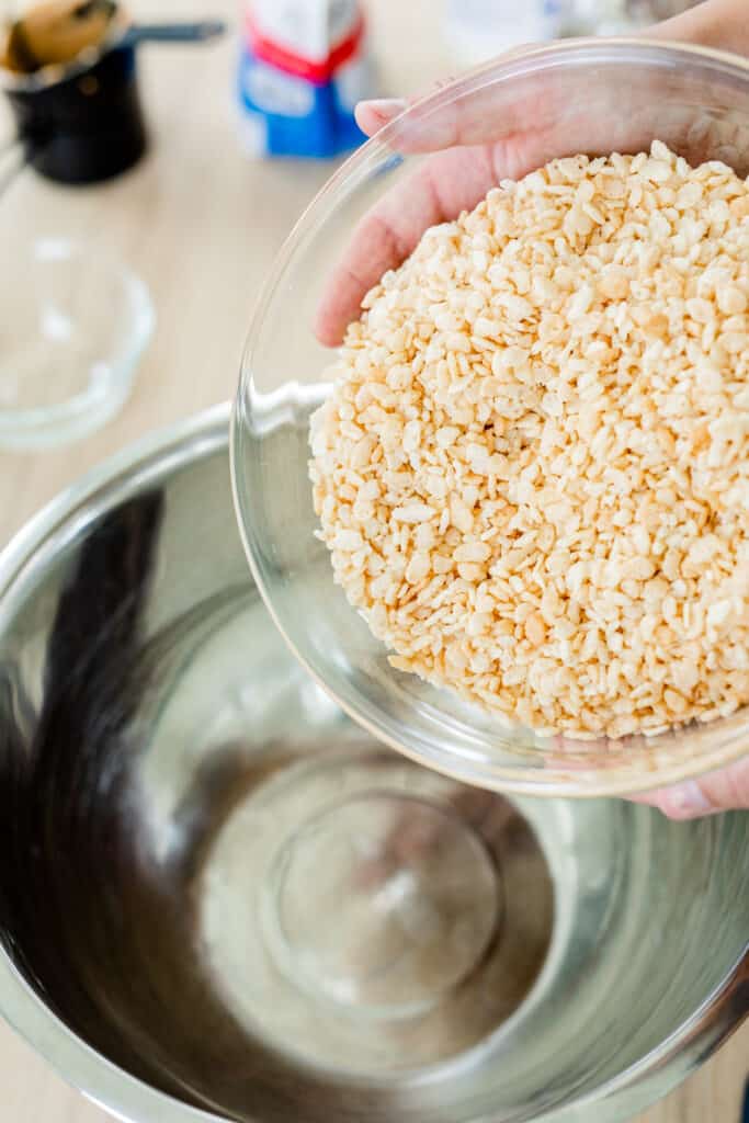 Rice Crispy cereals in a glass bowl are being poured into a large metal bowl.