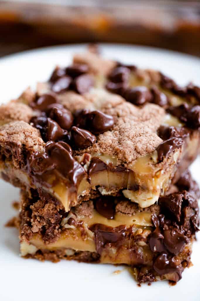 Two slices of german chocolate caramel brownies sit stacked on a white plate. A bite is taken from the corner of the brownie showing a thick layer of chewy caramel.