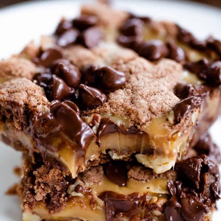 Two slices of brownie squares sit stacked on a white plate. A bite is taken from the corner of the brownie showing a thick layer of chewy caramel.