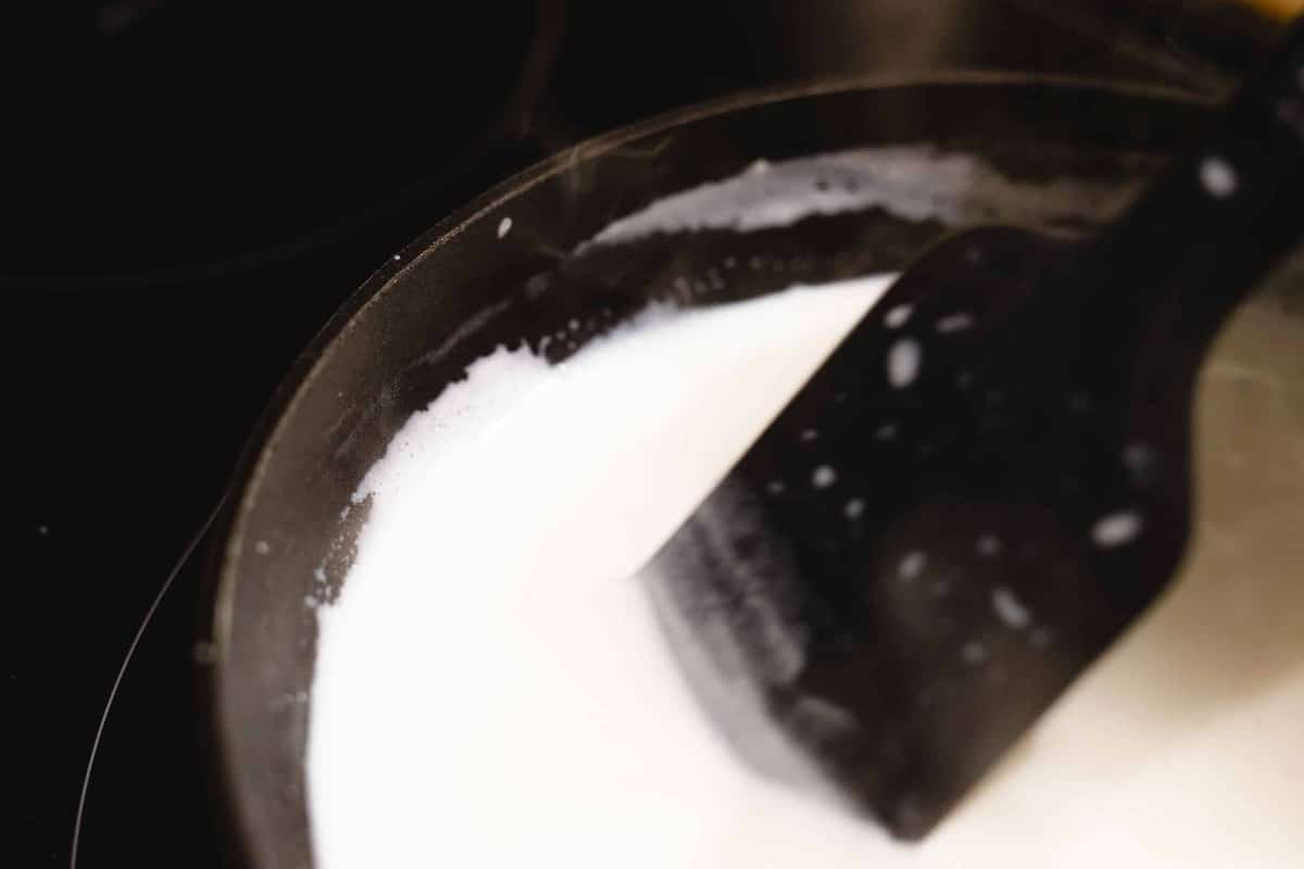A black spatula stirs milk gently as it heats up in a pot over the stove.