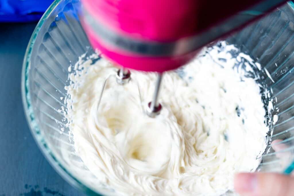 A hand mixer beats a block of cream cheese in a glass bowl.