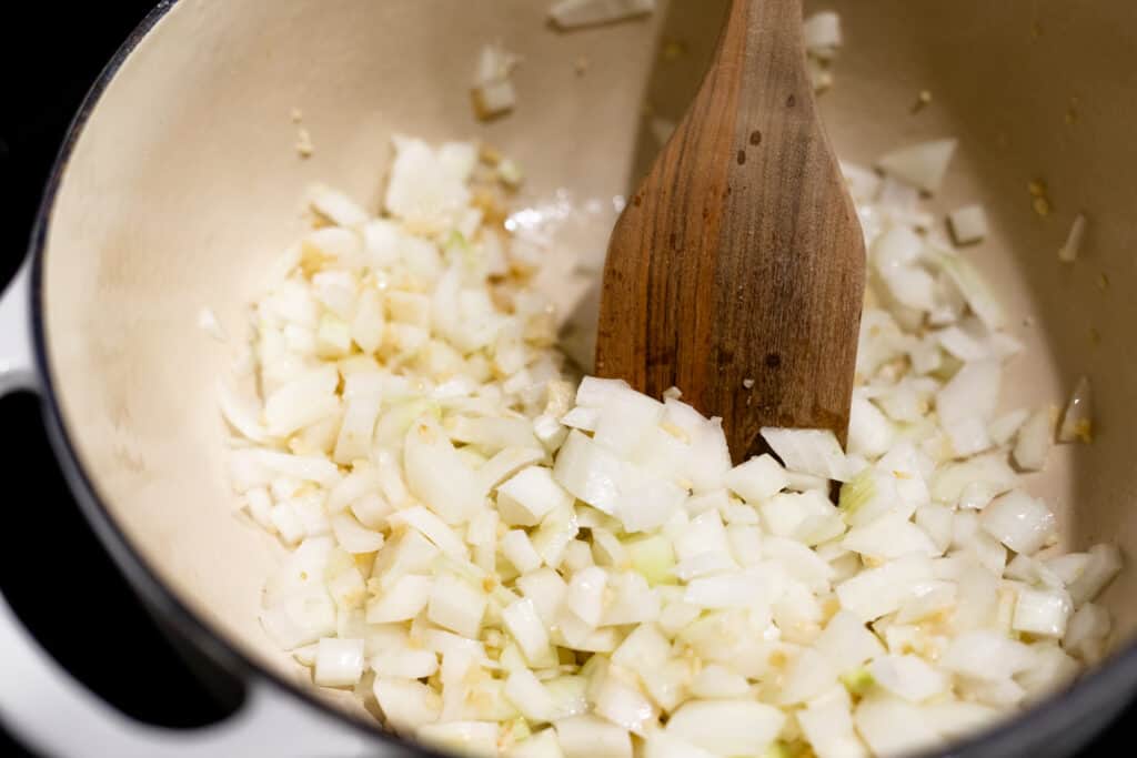 Diced onions, garlic and ginger paste are combined with a wooden spoon in a large pot.
