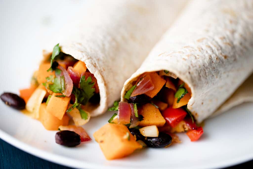 Black Bean Sweet Potato Burritos sit on a plate overfilled with potatoes, beans, cilantro, onion, peppers and chilis.