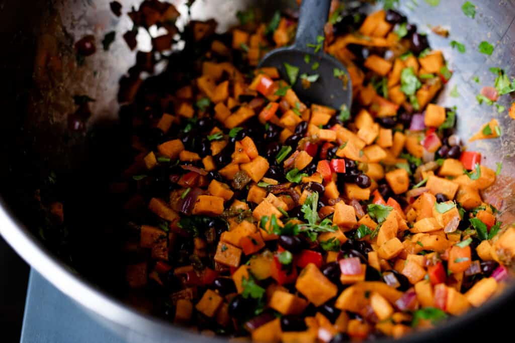 A metal bowl is filled with sweet potato, black beans, cilantro, peppers, chilis and is stirred by a spatula.