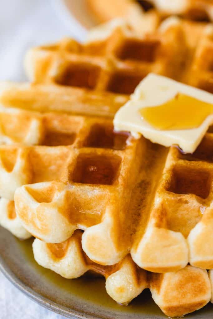 Two buttermilk waffles sit on a plate topped with a square tab of butter and rich homeamde syrup.