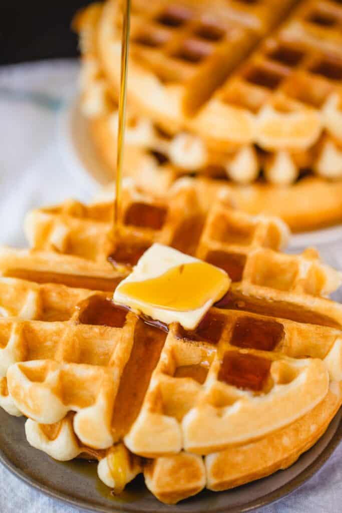 Golden brown buttermilk waffles sit on a plate, topped with butter and syrup they are ready to be enjoyed.