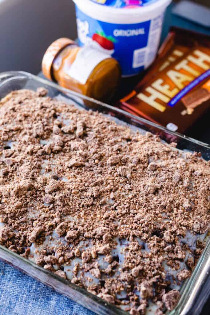 A cake covered in heath candy bits sits alongside the ingredients that make up the next layers of the cake, caramel, cool whip and more heath candy.