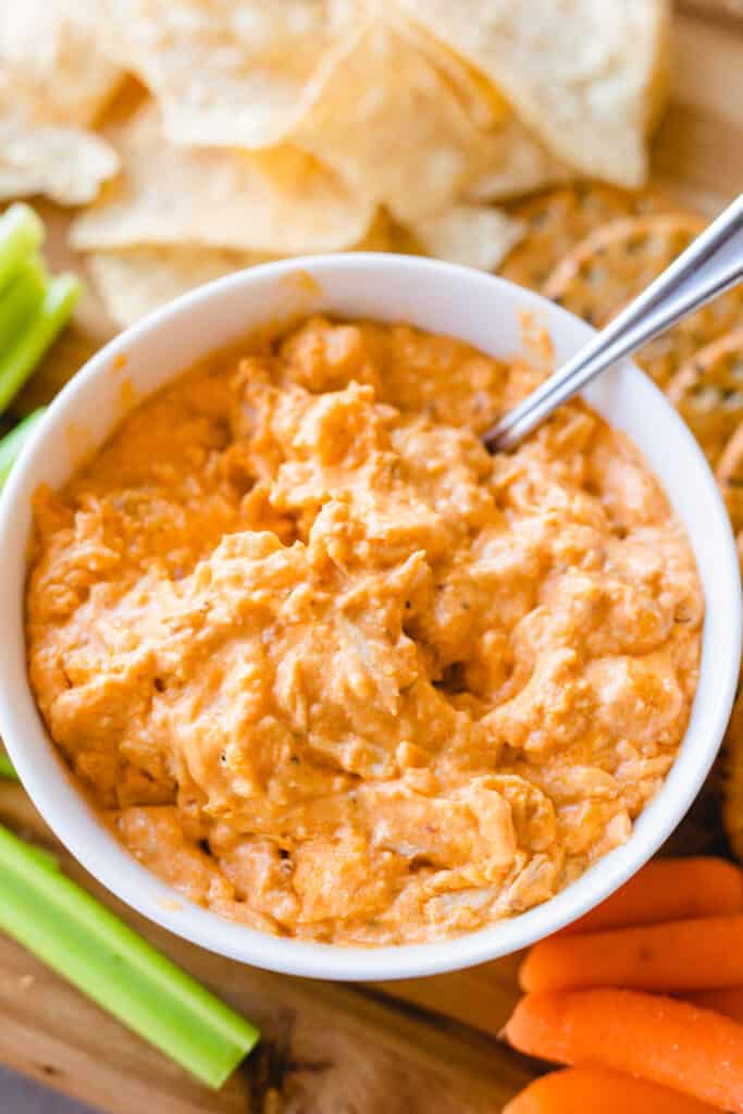 A white bowl filled with baked buffalo chicken dip sits surrounded by celery, tortilla chips, carrots and crackers for dipping.
