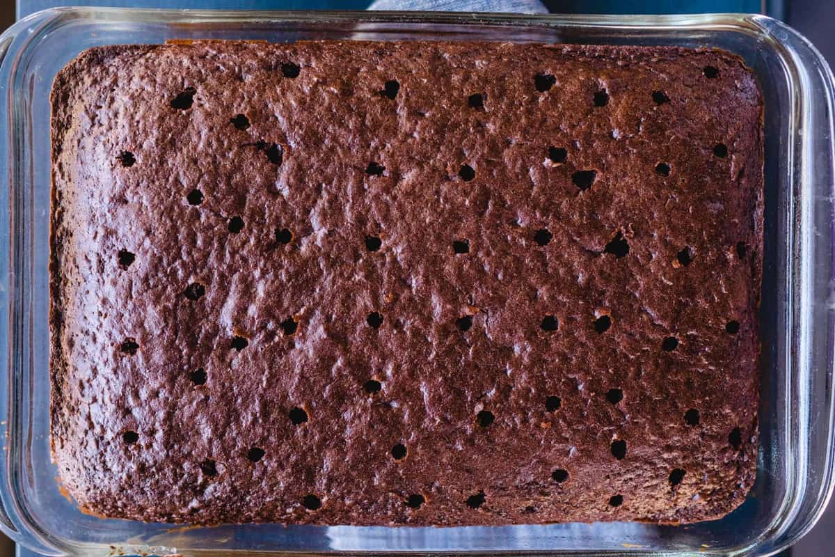 A warm baked chocolate cake in a 9x13 dish has holes poked throughout the top.
