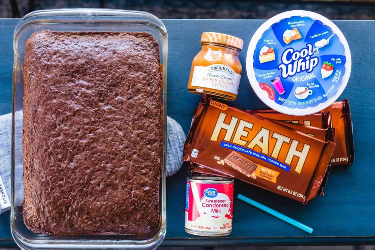 A chocolate cake, can of milk, jar of caramel sauce, tub of cool whip, heath candy bars sit on a counter top ready to assemble a cake.