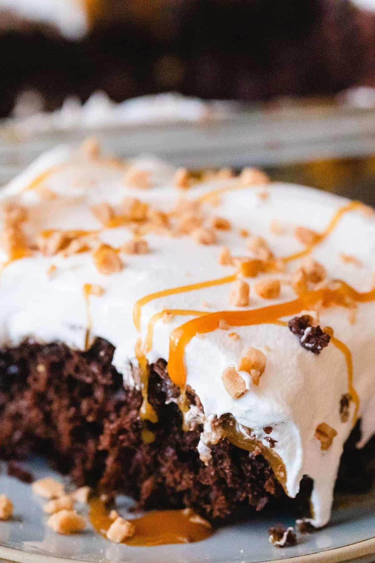 Square slice of chocolate cake sits on a plate. Topped with cool whip, toffee bits and drizzled caramel it is ready to eat.