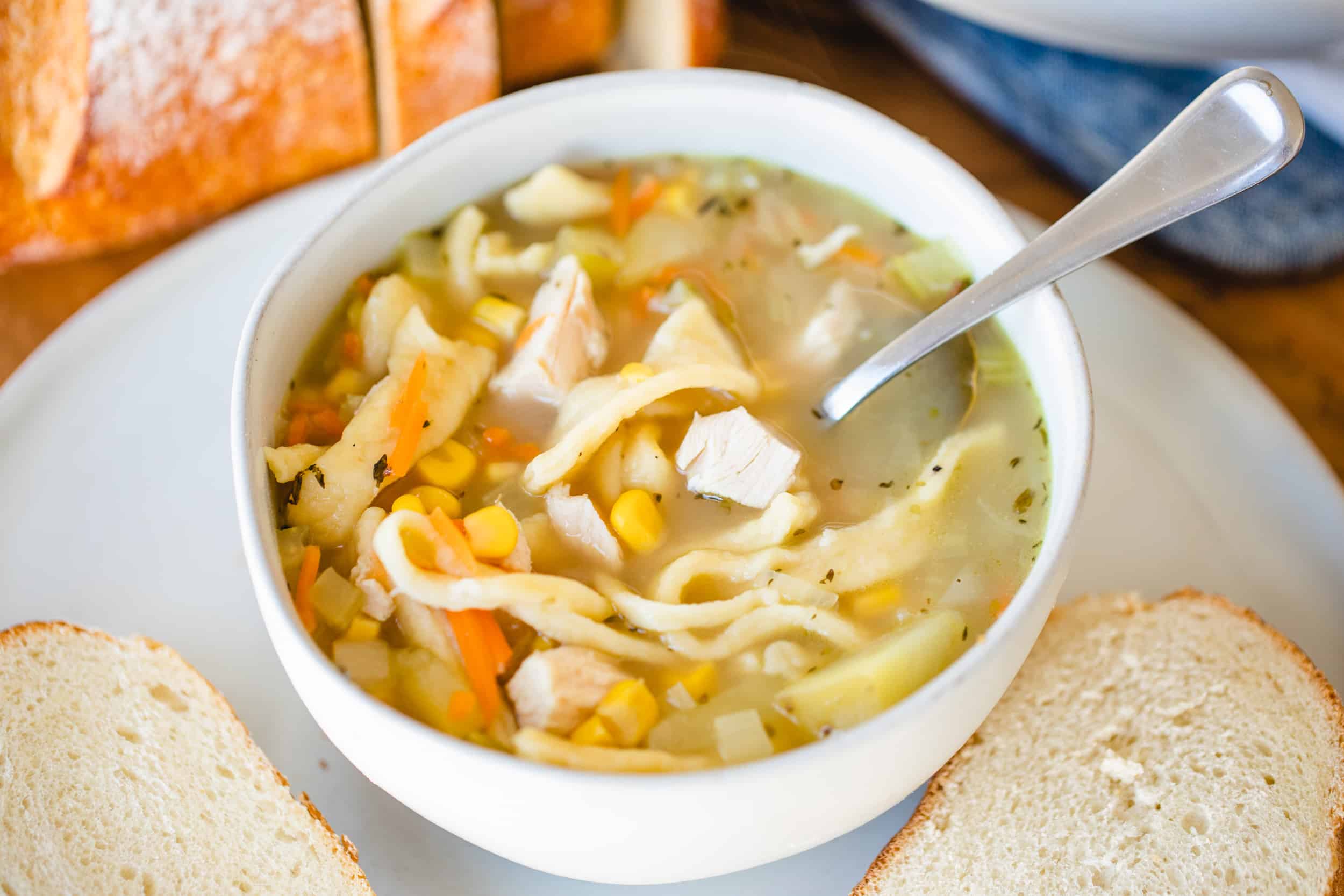 Bowl and spoon of chicken noodle soup sit beside sliced bread.