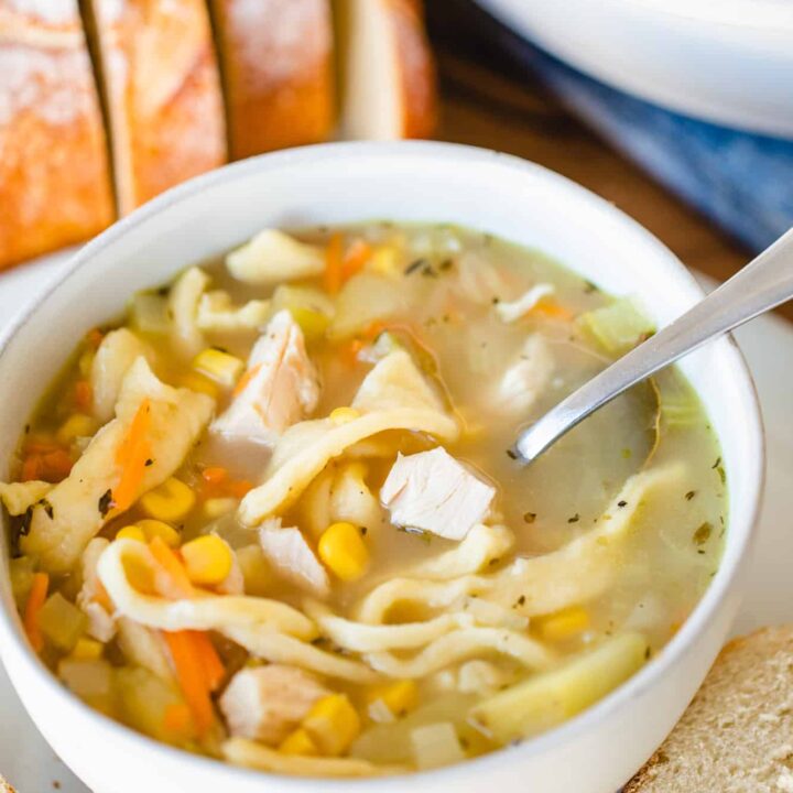 Bowl and spoon of chicken noodle soup sit beside sliced bread.