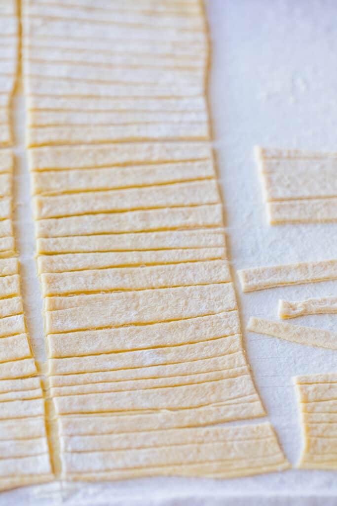 Strips of fresh cut homemade egg noodles sit on a floured bread cloth.