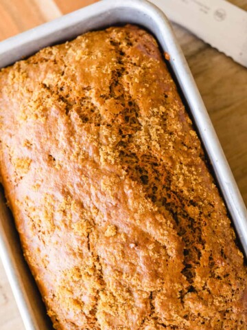 Pan of baked banana bread sits, cooling on a wooden cutting board alongside a large bread knife for slicing.