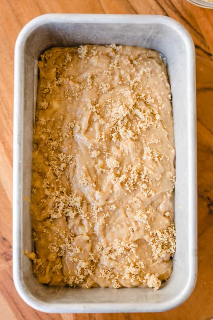 Brown sugar is crumbled on top of bread batter in a silver loaf pan.