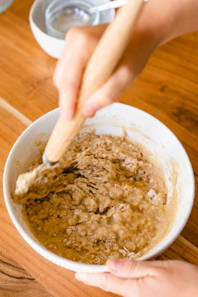 Ashley stirs banana bread batter with a dough whisk in a large white bowl.