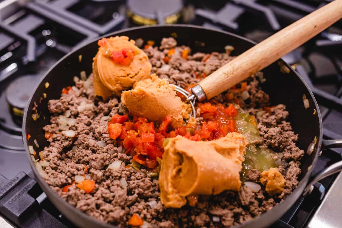 Cooked ground beef, onions and peppers are covered in enchilada saace, rotel, and refried beans sit in a pan over the stove.