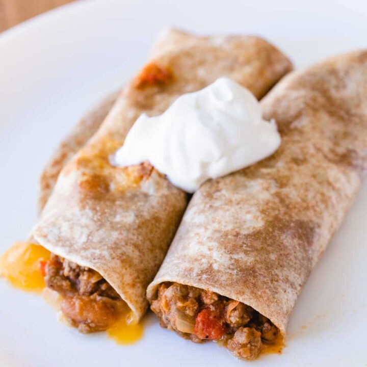 Two beef burritos sit on a white plate, topped with dollop of sourcream.