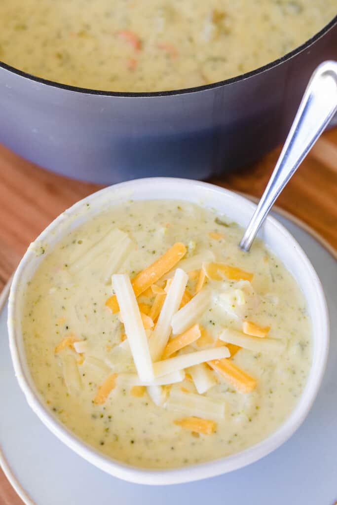 Bowl of cheddar broccoli soup is topped with shredded cheese and pot of soup behind it.