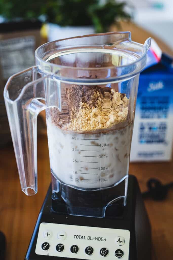 Almond milk, ice, water, coffee, protein and cacao powder sit in a blender on a wooden table top.