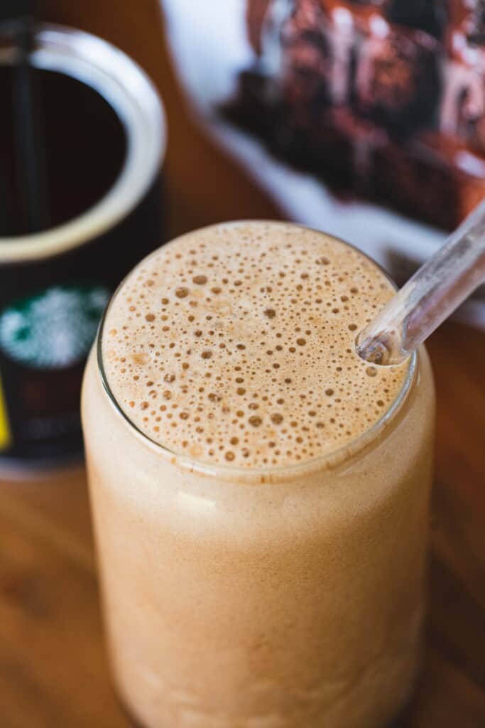 A clear tall glass is filled to the brim with peanut butter chocolate mocha protein shake with a clear straw.