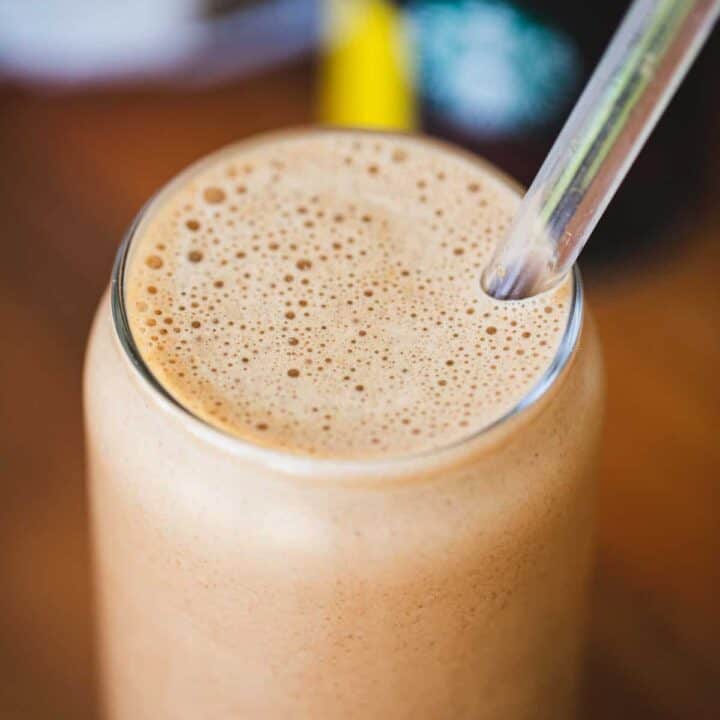 A clear tall glass is filled to the brim with peanut butter chocolate mocha protein shake with a clear straw.