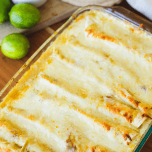 Wooden spatula sits in a pan of cheesy enchiladas. A serving of enchiladas sits on a plate beside the pan.