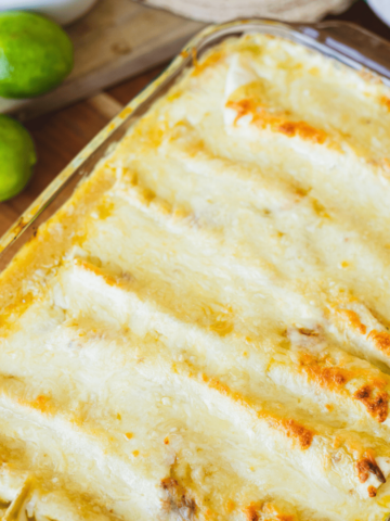 Wooden spatula sits in a pan of cheesy enchiladas. A serving of enchiladas sits on a plate beside the pan.