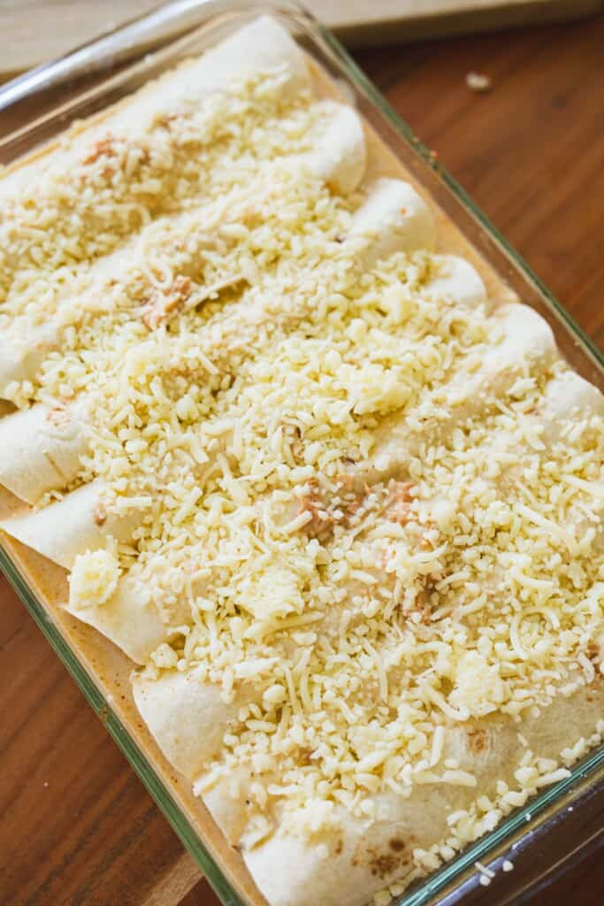 Shredded cheese is added to the top of a pan of prepared enchiladas and are ready to bake.