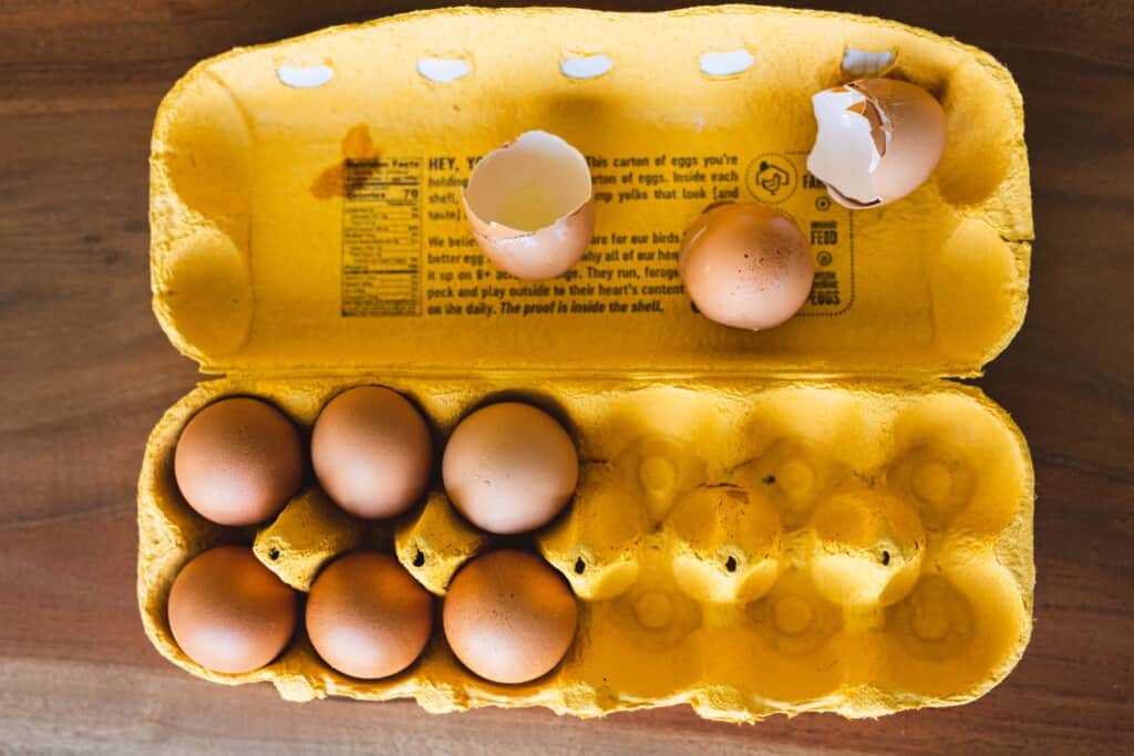 A yellow egg carton with six eggs sits on a counter. Three cracked egg shells sit in the box.