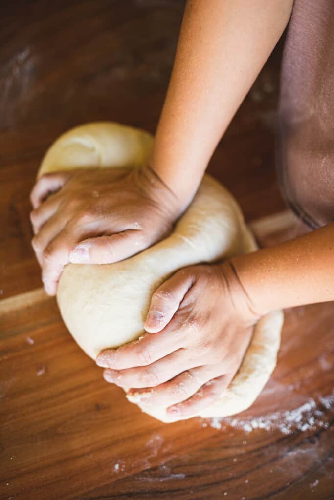 Ashley kneads a loaf of dough with her hands on a wooden countertop.
