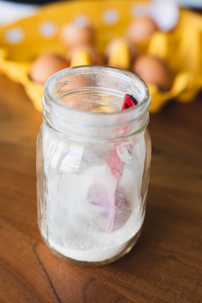A glass mason jar of salt with a red measuring spoon sits on a wooden counter top in front of an open egg carton.