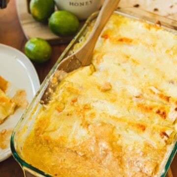 Wooden spatula sits in a pan of cheesy enchiladas. A serving of enchiladas sits on a plate beside the pan.