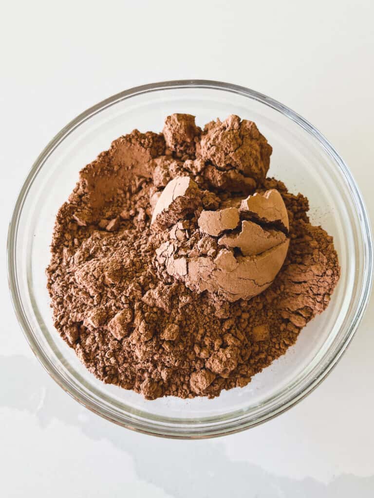 Scoops of cacao powder sit in a glass bowl on a white countertop.
