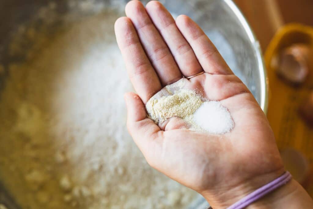 Ashley holds measured out salt and dough enhancer in her open palm over a metal bowl of ingredients.
