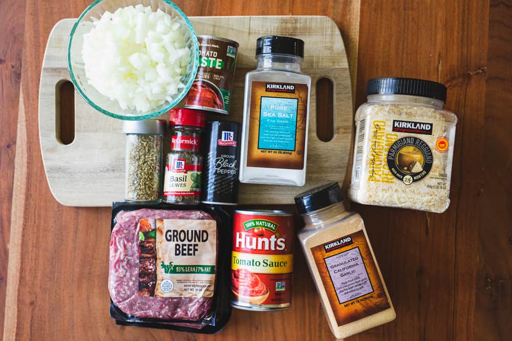 Ingredients for spaghetti sauce sit on a wooden countertop with a worn wooden cutting board.