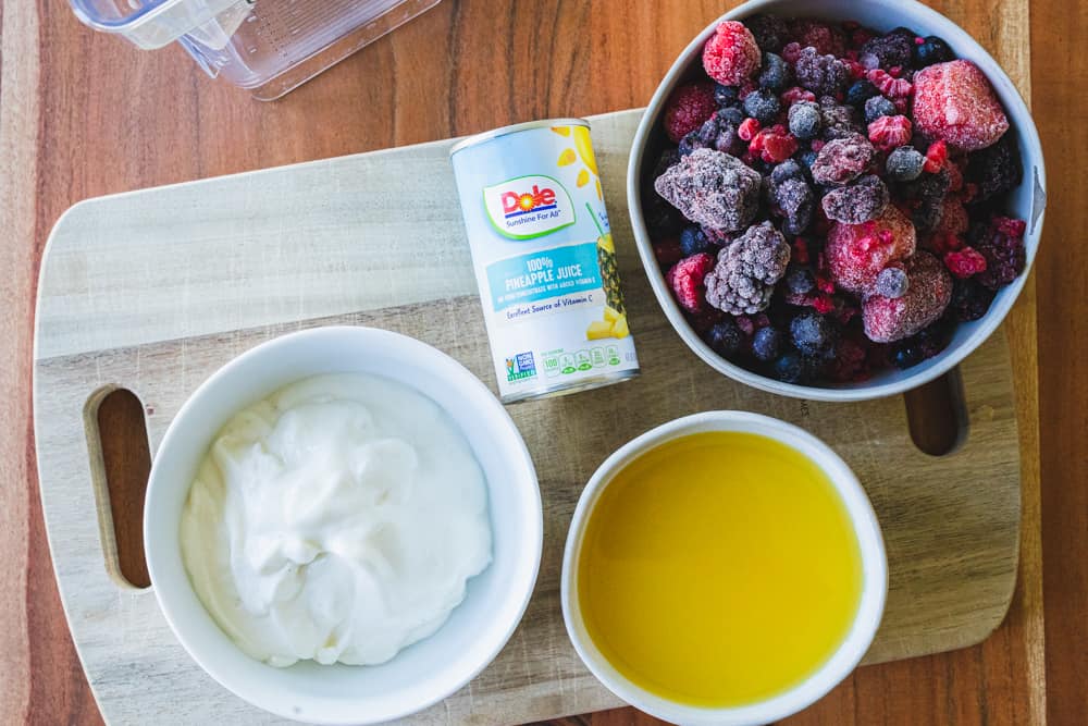 Bowls of yogurt, juice, frozen berries and a can of pineapple juice sit on a wooden cutting board.