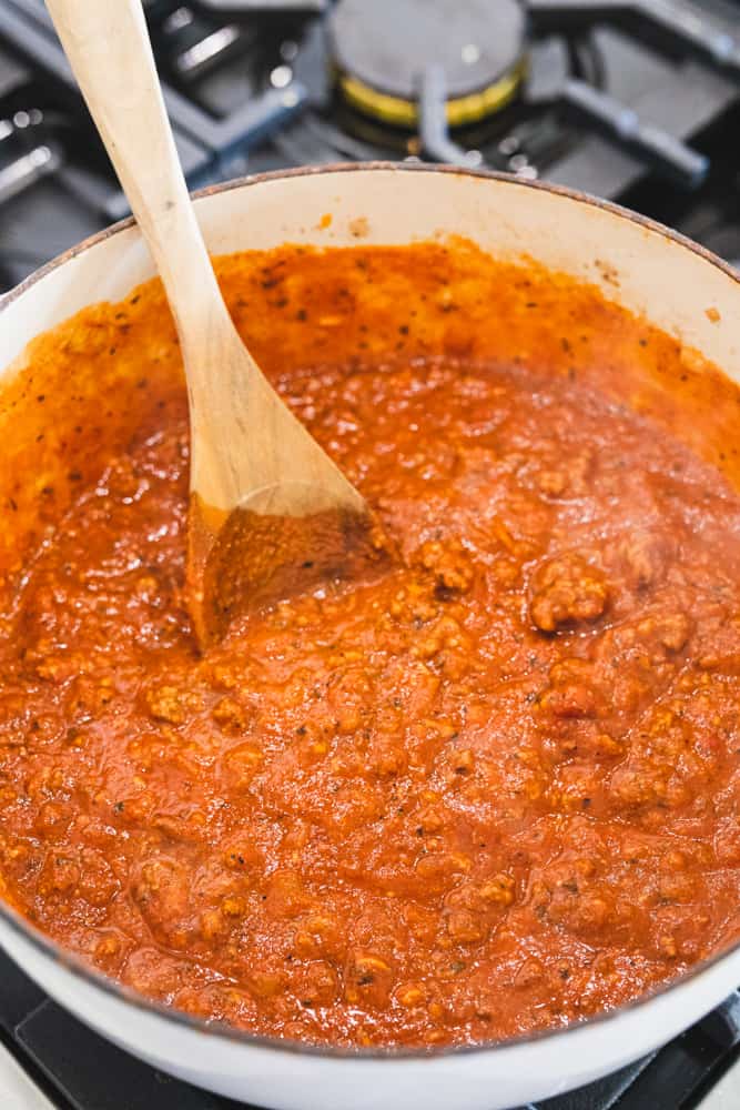 Wooden spoon stirs stewing spaghetti meat sauce in a large pot over the stove top.