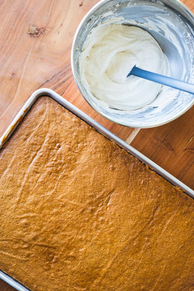 Bowl of cinnamon cream cheese frosting sits beside a cooled pan of sheet cake.