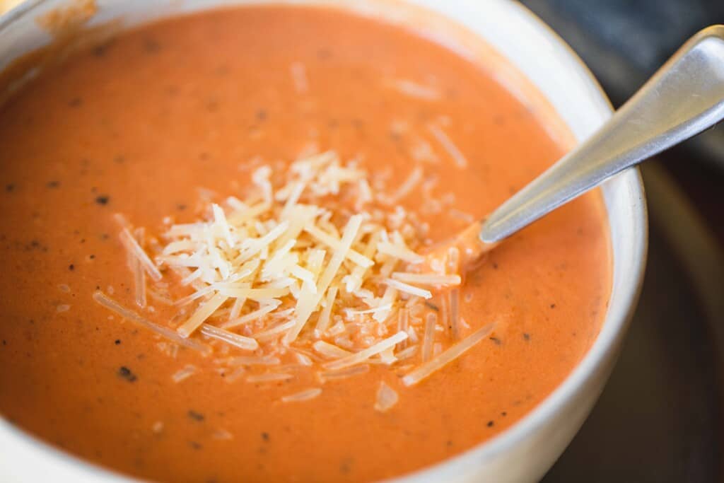 A white ceramic bowl filled with easy creamy tomato soup is topped with shredded parmesan cheese with a silver spoon.