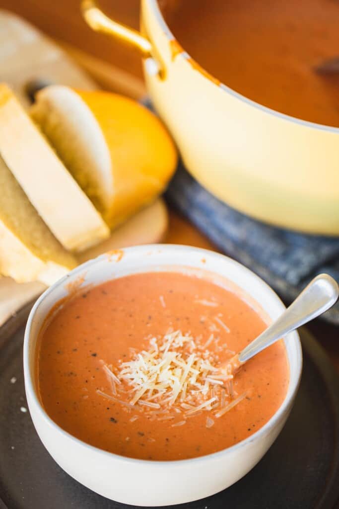 Bowl of creamy tomato soup topped with parmesan sits in front of a thick sliced french bread loaf and large pot of soup.