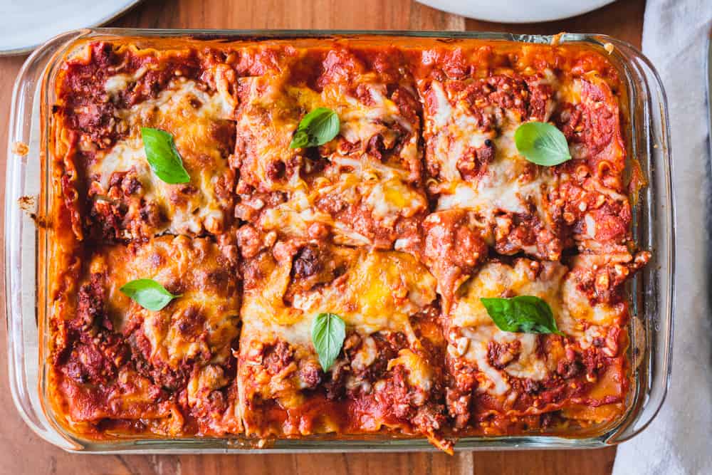 Pan of easy lasagna with cottage cheese sits on wooden counter sliced into 6 large sections. Each section is topped with a fresh green basil leaf.