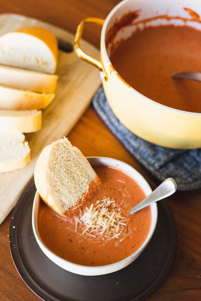 Bowl of tomato soup is served with a slice of french bread. Alongside a full pot of cooked tomato soup and loaf of sliced bread.